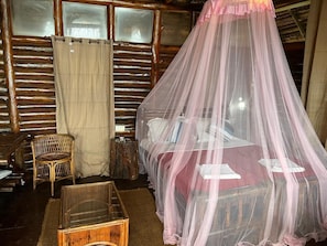 Una habitación rústica con una cama bajo un dosel rosa, una silla de mimbre y una mesita de noche de madera.