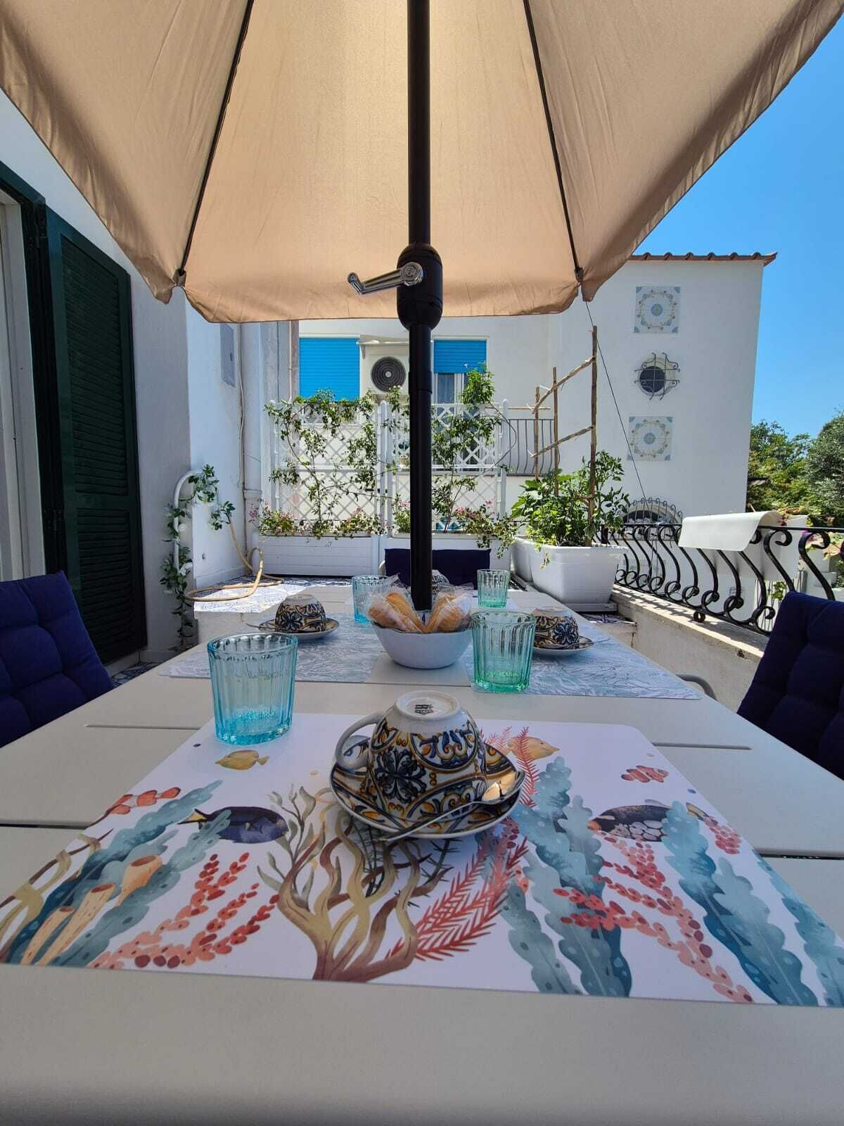 An outdoor dining area with a table set for two, a beige umbrella, and a view of a white building with blue shutters.