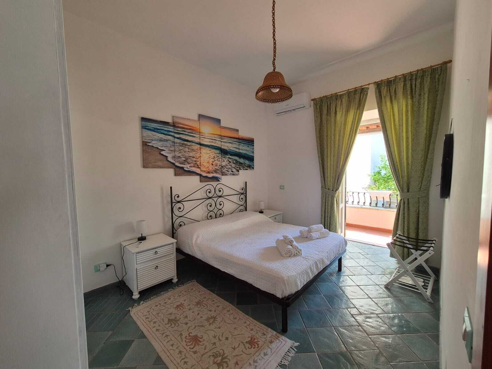 A bedroom with a bed, a nightstand, a wall-mounted air conditioner, a decorative painting, and a view of the outdoors through a window with curtains.