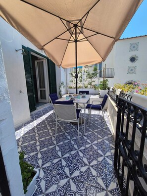 Eine Terrasse im Freien mit Tisch und Stühlen, einem beige farbenen Sonnenschirm und einem Fliesenboden mit Muster.