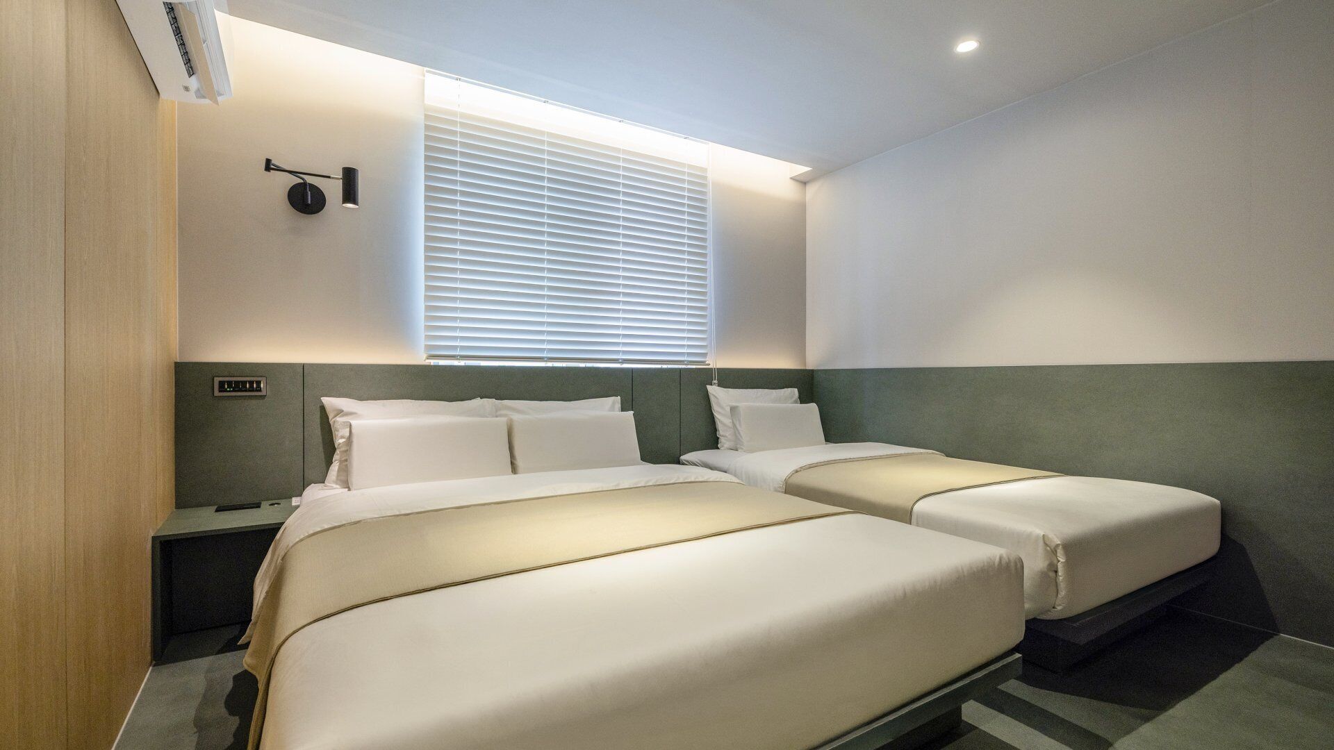 A hotel room with two beds, a bedside table, a lamp, and a window with blinds.