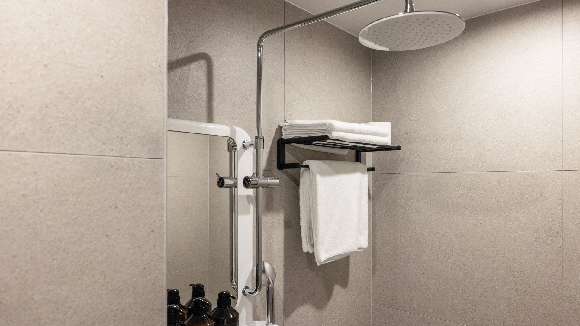 A shower with a handheld showerhead, a towel rack with folded towels, and a bottle of shampoo.