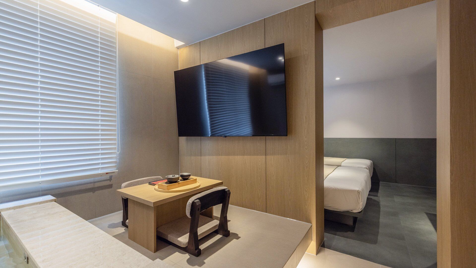 Una habitación de hotel moderna con una televisión de pantalla plana en la pared, un escritorio de madera con una silla, una mesita con cuencos y una cama con cabecera gris.