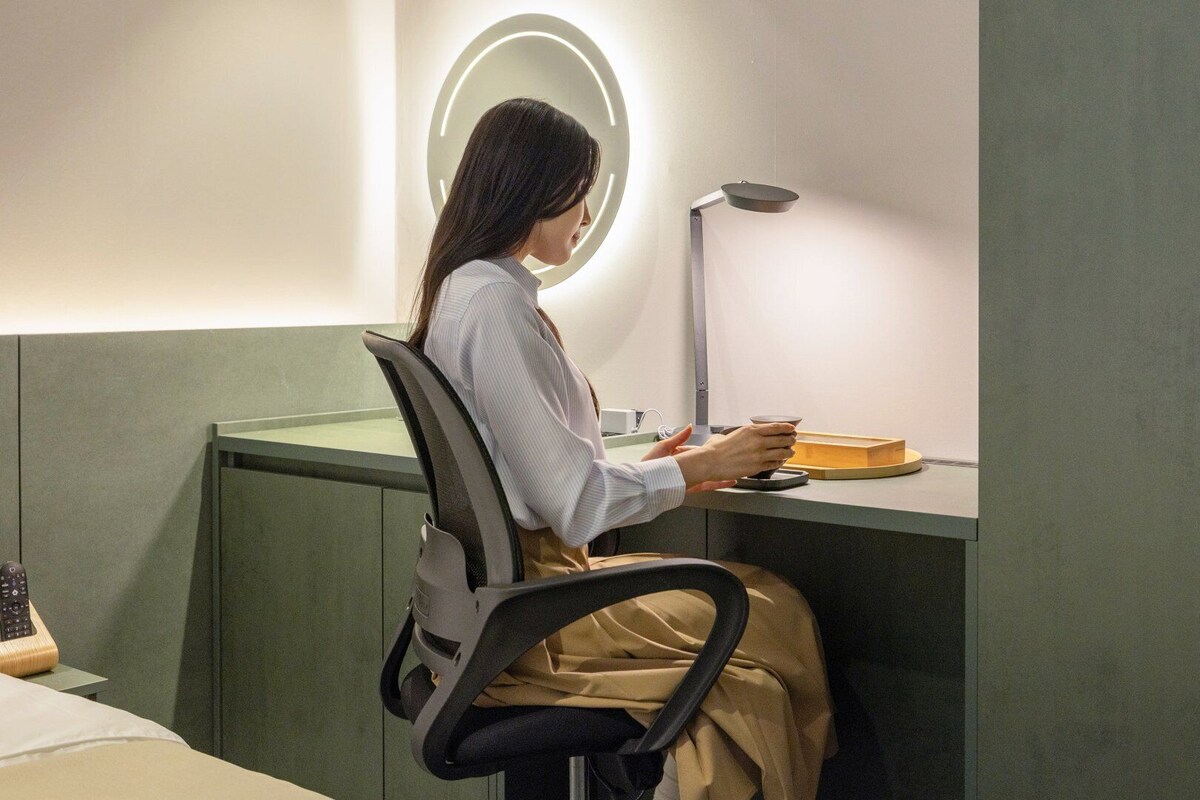 Una persona sentada en un escritorio, con una lámpara, una taza y una caja, en una habitación con un muro verde decorativo y una luminaria circular.
