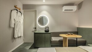 A hotel room with a bed, a bedside table with a lamp, a round mirror, a seating area with a table, and a coat rack with robes.