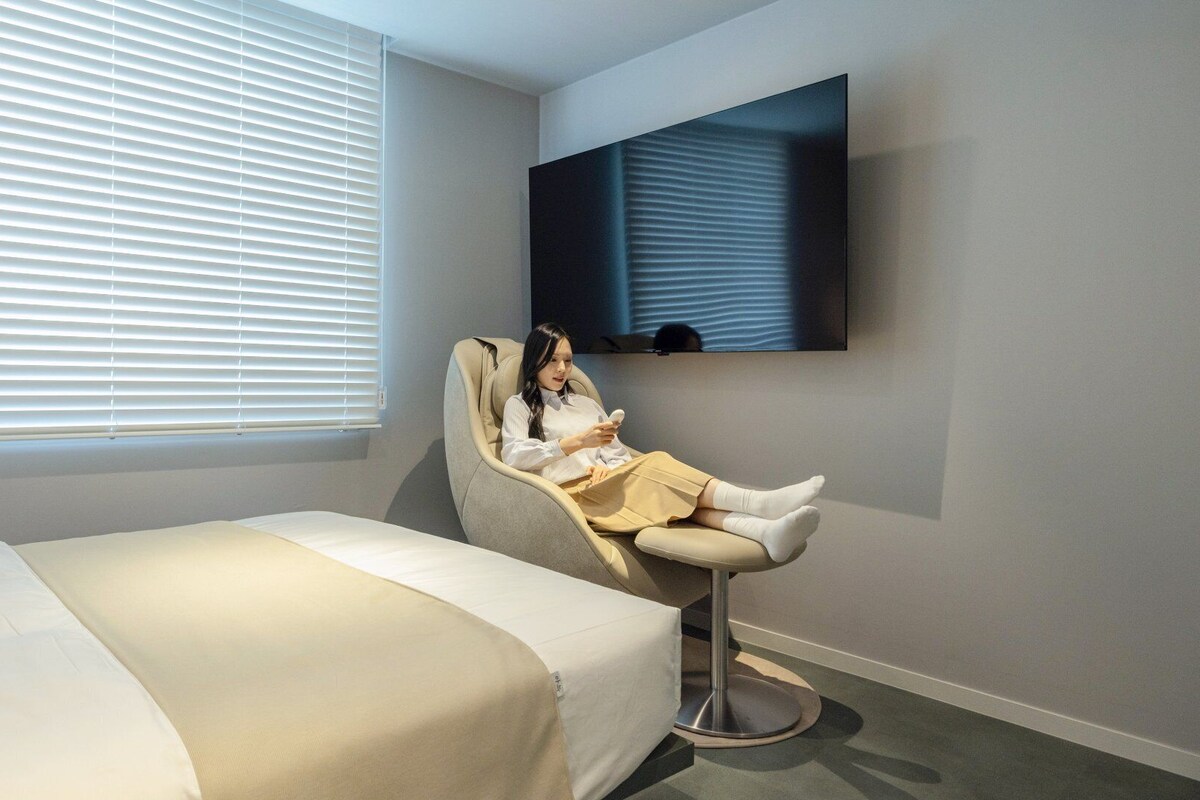 Habitación de hotel con una cama, una silla de masaje, un televisor montado en la pared y una ventana con persianas.