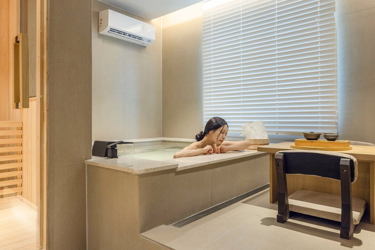 Un cuarto de spa con una bañera, una mesa de madera y una ventana con persianas.