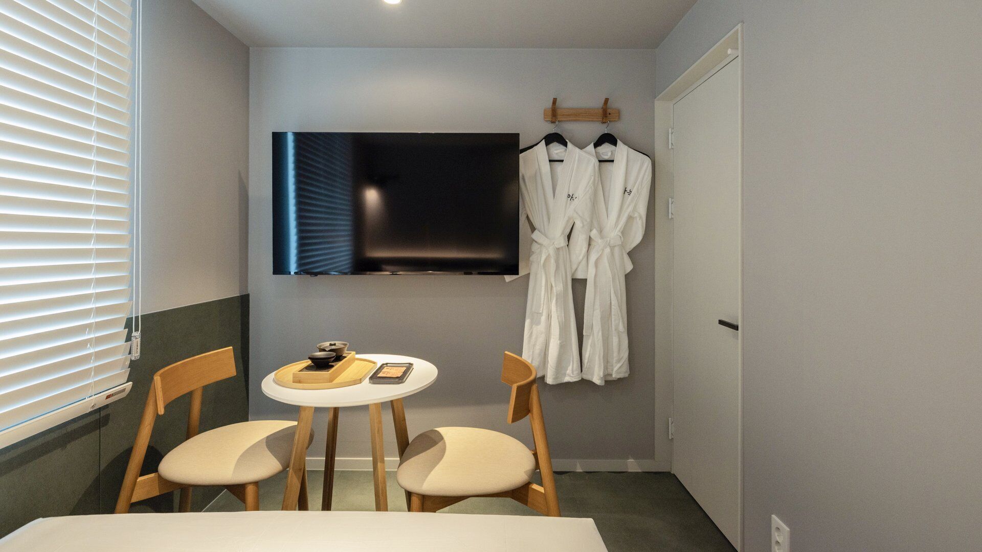 Una habitación de hotel pequeña con un televisor montado en la pared, una mesa con sillas y dos batas blancas colgadas en un perchero.