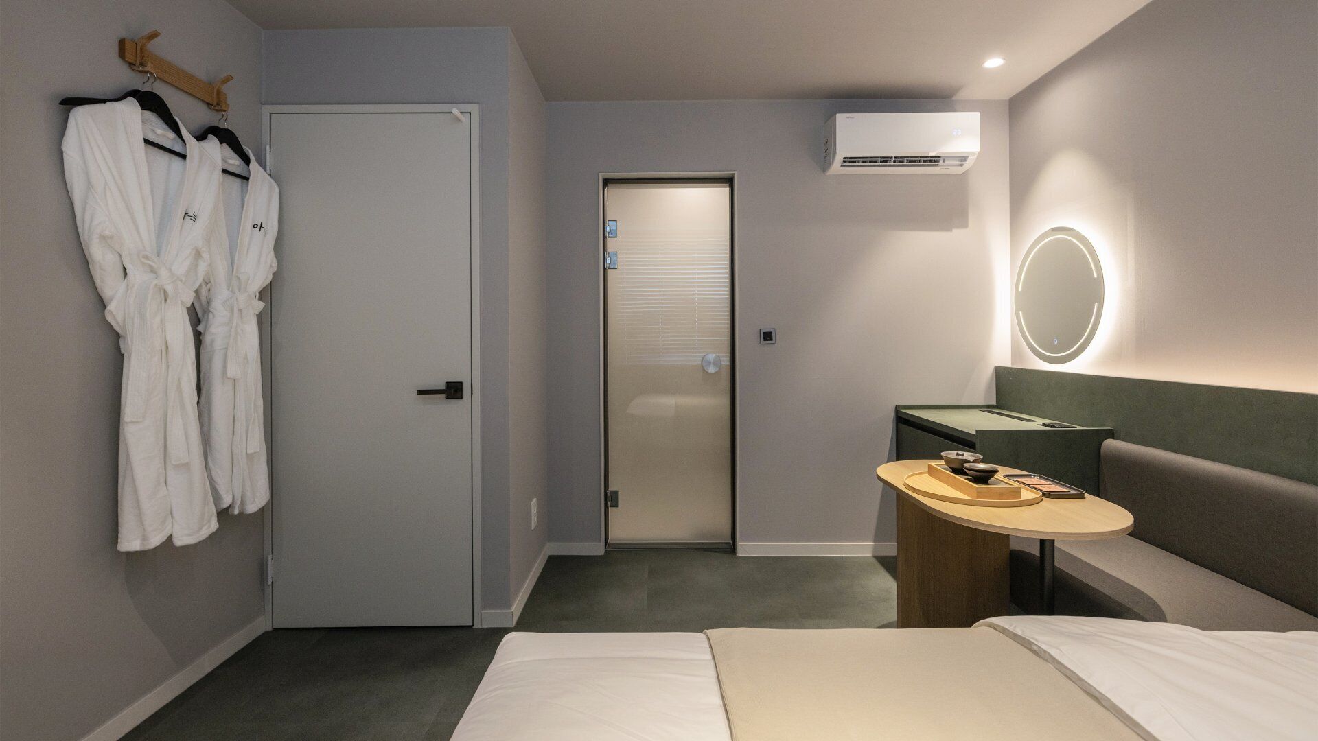 Habitación de hotel con cama, mesita de noche, espejo, perchero para bata y puerta que da al baño.