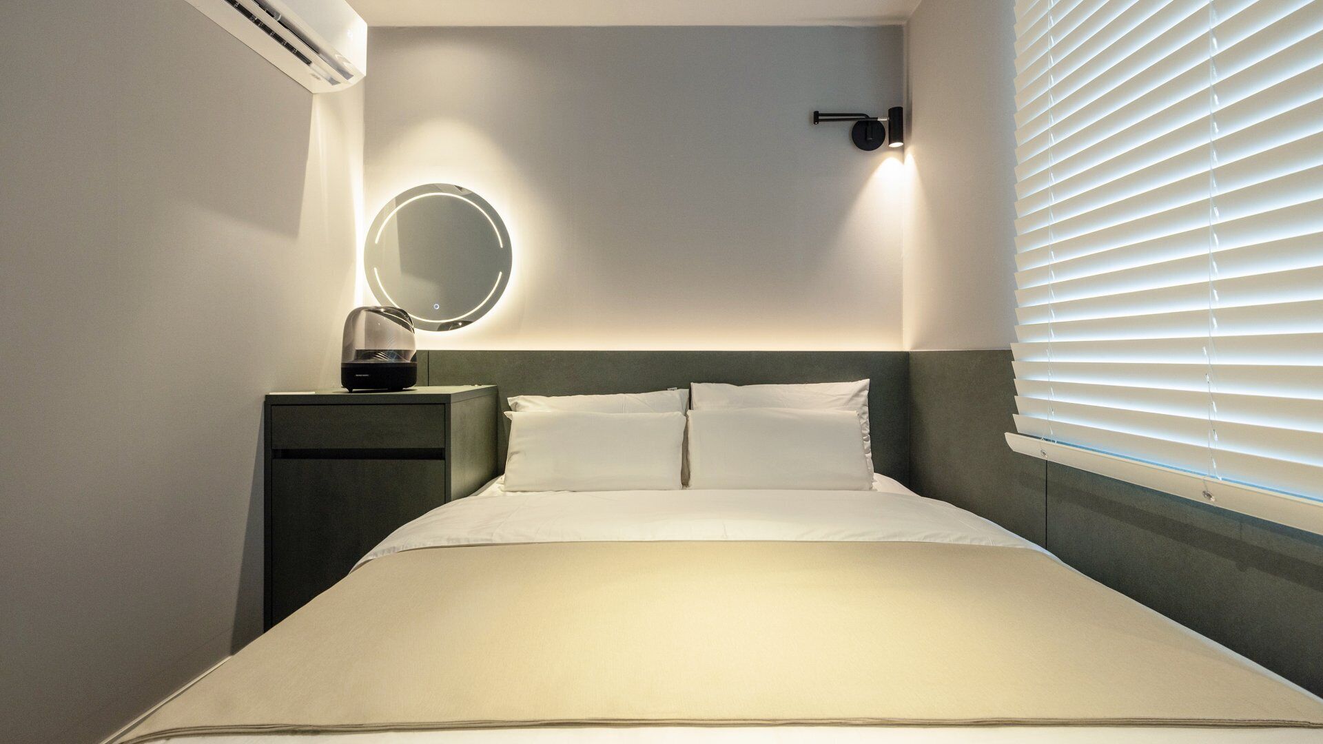 Habitación de hotel con cama, mesita de noche con lámpara, aire acondicionado instalado en la pared y ventana con persianas.