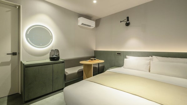 Habitación de hotel con cama, mesita de noche, espejo redondo, unidad de aire acondicionado y lámpara en la pared.