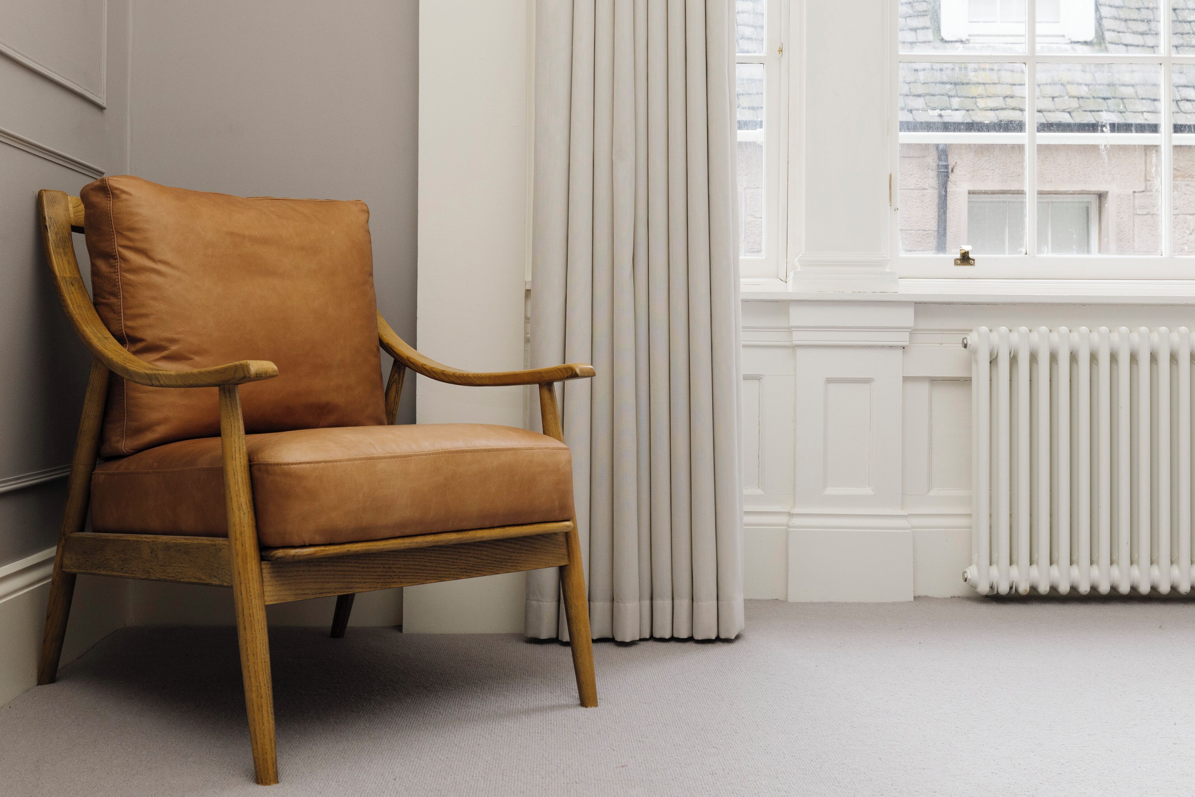 A wooden armchair with a cushion in a room with a radiator, a window with curtains, and a plain wall.