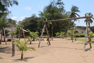 A wooden swing set in a sandy outdoor area with palm trees and a building in the background.