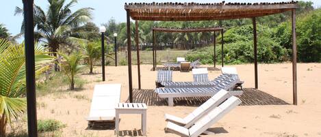 Un área de descanso frente a la playa con tumbonas, una mesita y una pérgola que ofrece sombra.