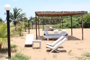 Un área de descanso frente a la playa con tumbonas, una mesita y una pérgola que ofrece sombra.