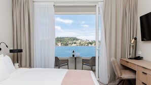 A hotel room with a bed, a desk, a chair, a TV, and a view of the sea and a hillside. - Hotel M (ZATON)