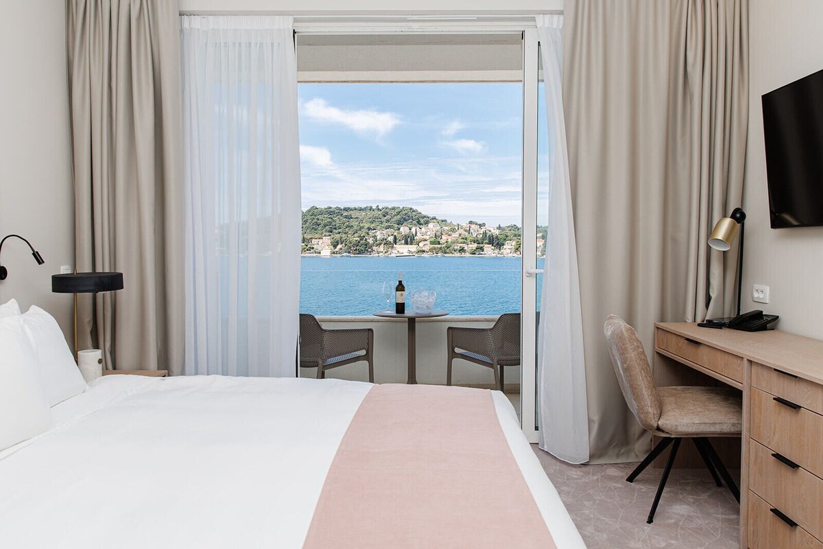 Ein Hotelzimmer mit Bett, Schreibtisch, Stuhl, Fernseher und Blick auf das Meer und eine Hügellandschaft.