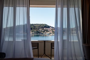 A room with a view of a lake and a hillside town through sheer curtains.