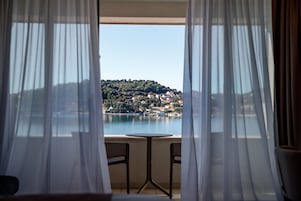 A room with a view of a lake and a hillside town through sheer curtains.