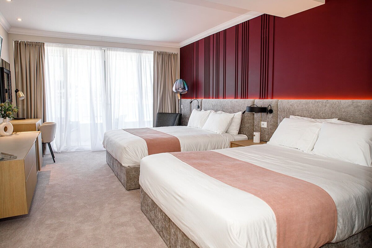Ein Hotelzimmer mit zwei Betten, einem Schreibtisch, einem Stuhl und einem großen Fenster mit Vorhängen.
