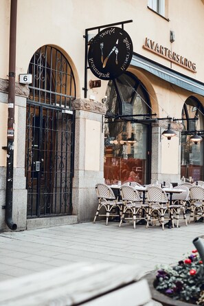 Ein Gebäude mit einem Schild, auf dem 'Kvarterskrog' steht, und einer schwarzen Uhr mit Besteck. Vor dem Gebäude befinden sich Tische und Stühle im Freien.