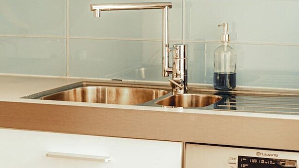 Una cocina con un lavamanos de acero inoxidable, lavavajillas y un reloj digital en la pared.