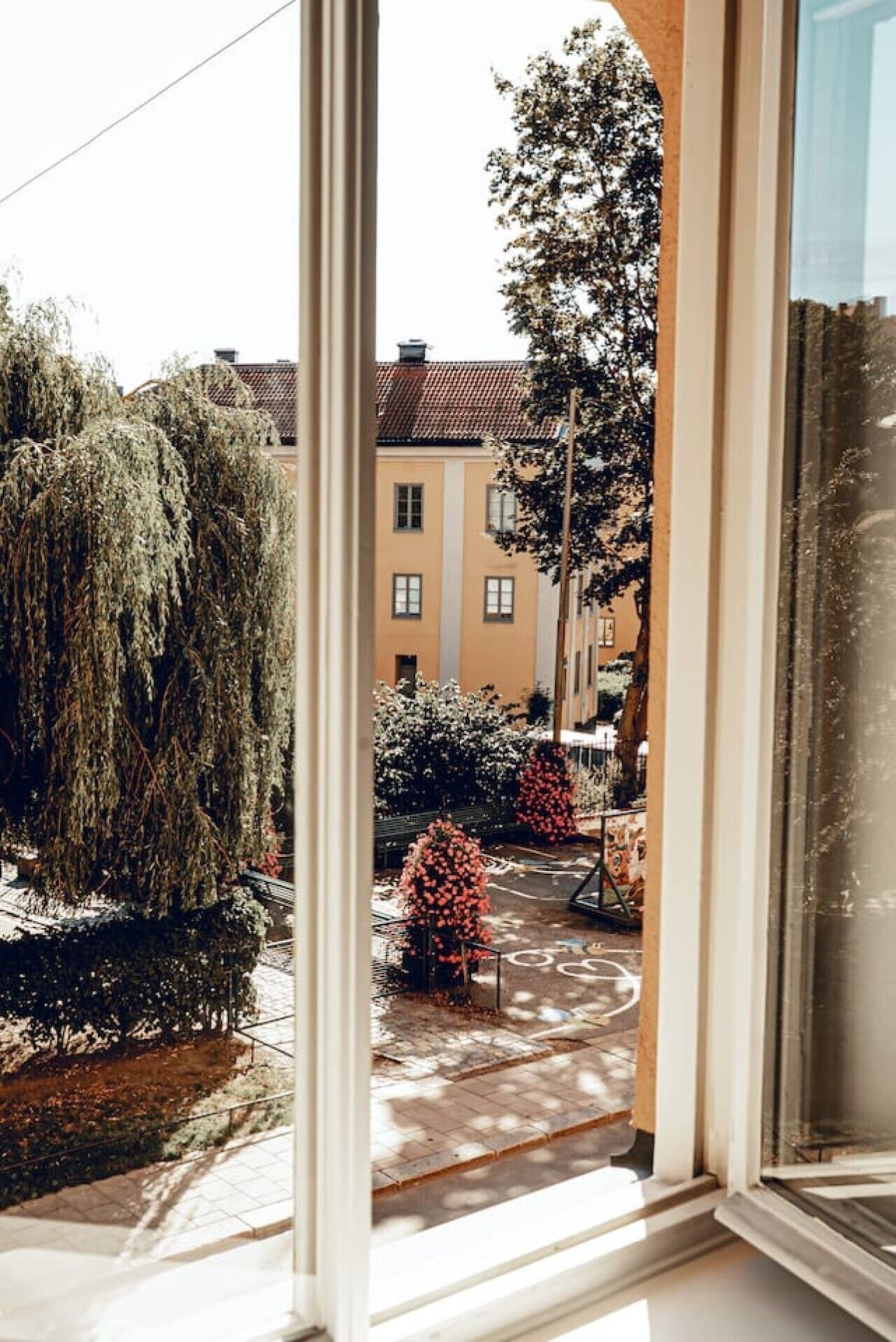 A view through a window of a courtyard with trees, a bench, and buildings in the background.