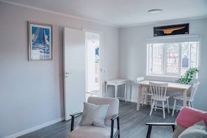 A room with a dining table, chairs, a sofa, a window with a view, and a door leading outside. - Royal Yacht Club Apartment — Sandhamn Harbour (Sandhamn)