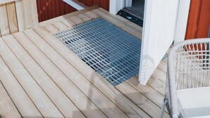 Puinen terassi, jonka sisäänkäynnissä on metallinen ritilä, valkoinen ovi ja pieni puinen rakennelma vasemmalla puolella.