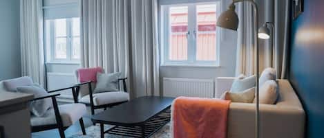 A modern living room with a sofa, chairs, a coffee table, and a floor lamp. There is a window with curtains and a radiator.