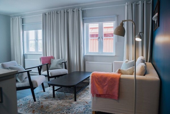 A modern living room with a sofa, chairs, a coffee table, and a floor lamp. There is a window with curtains and a radiator.