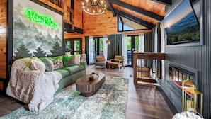 A cozy cabin interior with a green neon sign, a fireplace, and a large window overlooking a forest. - Velvet Pines | Lake Front | Amenities Galore | 5* (Pigeon Forge)