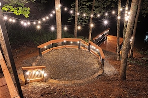 A circular outdoor seating area with a wooden bench and a small table, surrounded by trees and string lights. - Velvet Pines | Lake Front | Amenities Galore | 5* (Pigeon Forge)