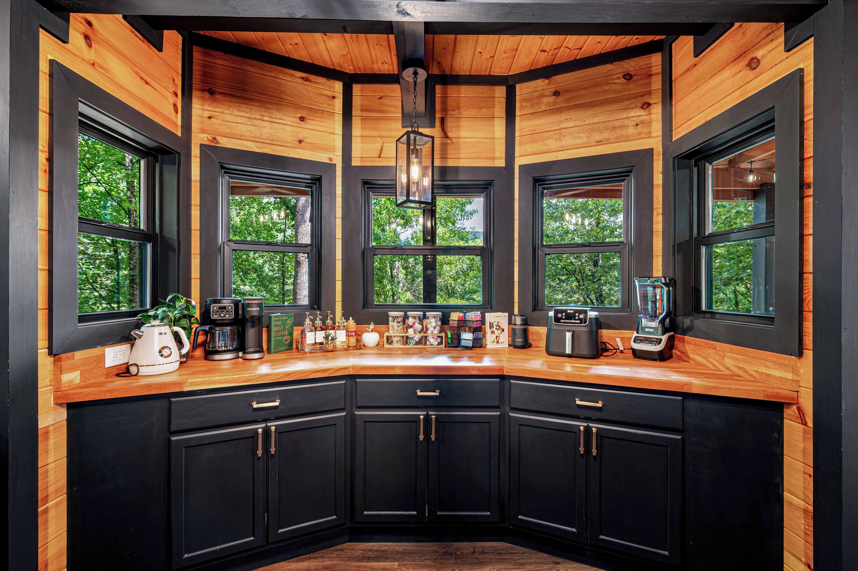 A modern kitchen with black cabinets, wooden countertops, and large windows offering a view of trees.