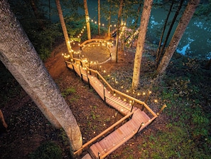 Un escalier en bois mène à un espace circulaire aménagé en coin salon, entouré d’arbres, et des guirlandes lumineuses sont disposées le long du chemin.