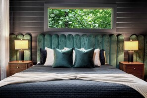 Quarto com cama grande, duas mesas de cabeceira, janela com vista para vegetação e cabeceira estofada verde.