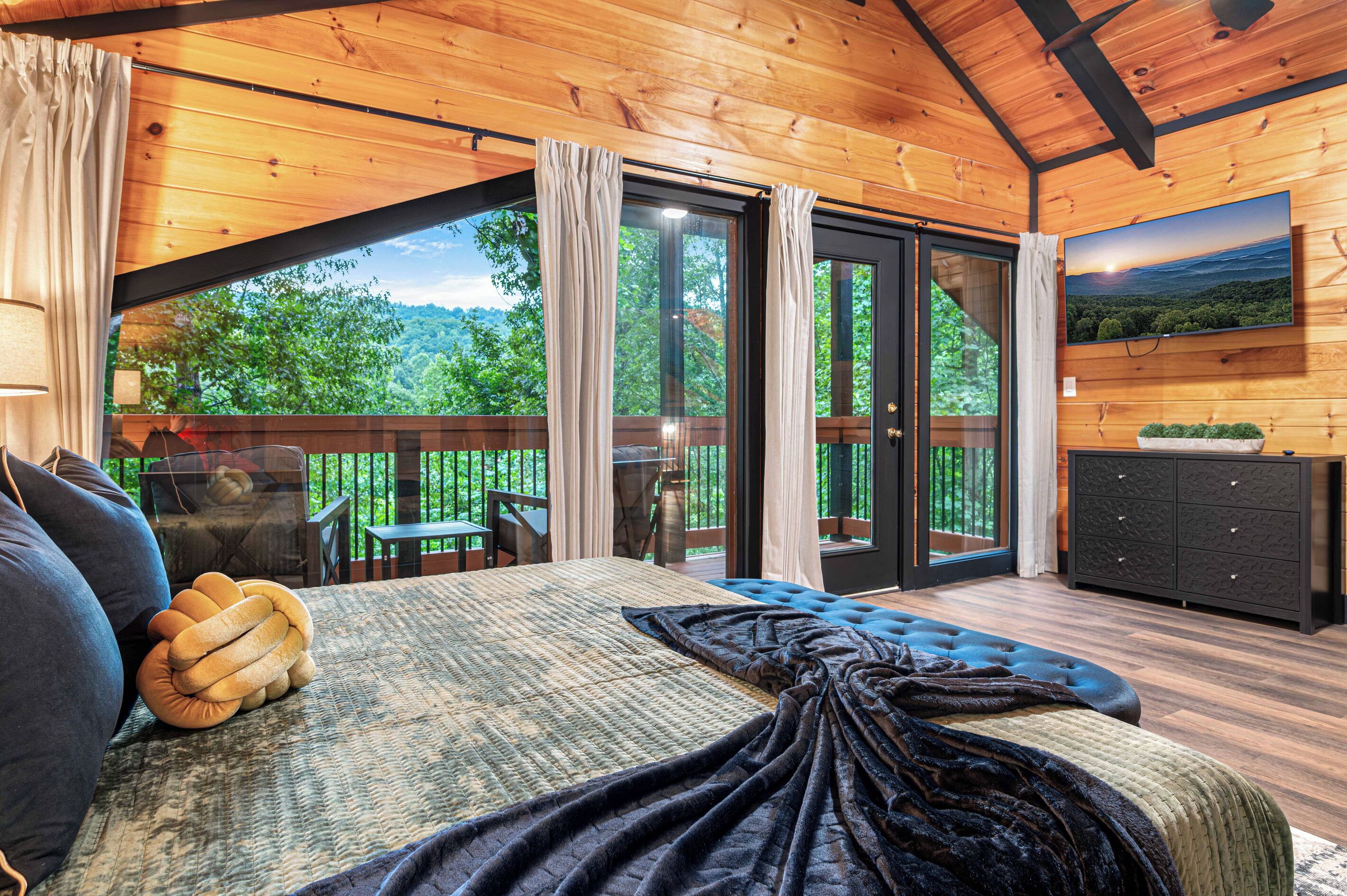 A bedroom with a large bed, a view of the outdoors, a dining area, and a TV on the wall.