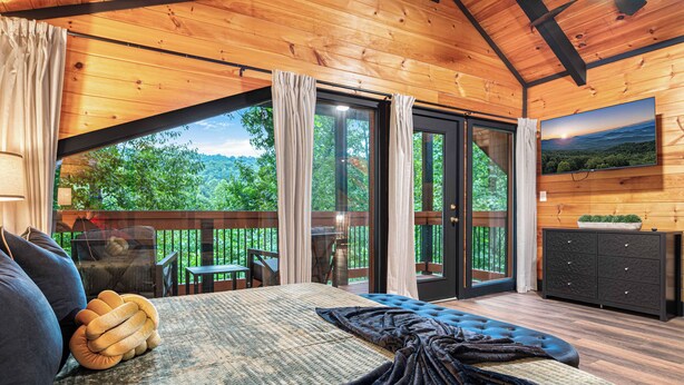 A bedroom with a large bed, a view of the outdoors, a dining area, and a TV on the wall. - Velvet Pines | Lake Front | Amenities Galore | 5* (Pigeon Forge)
