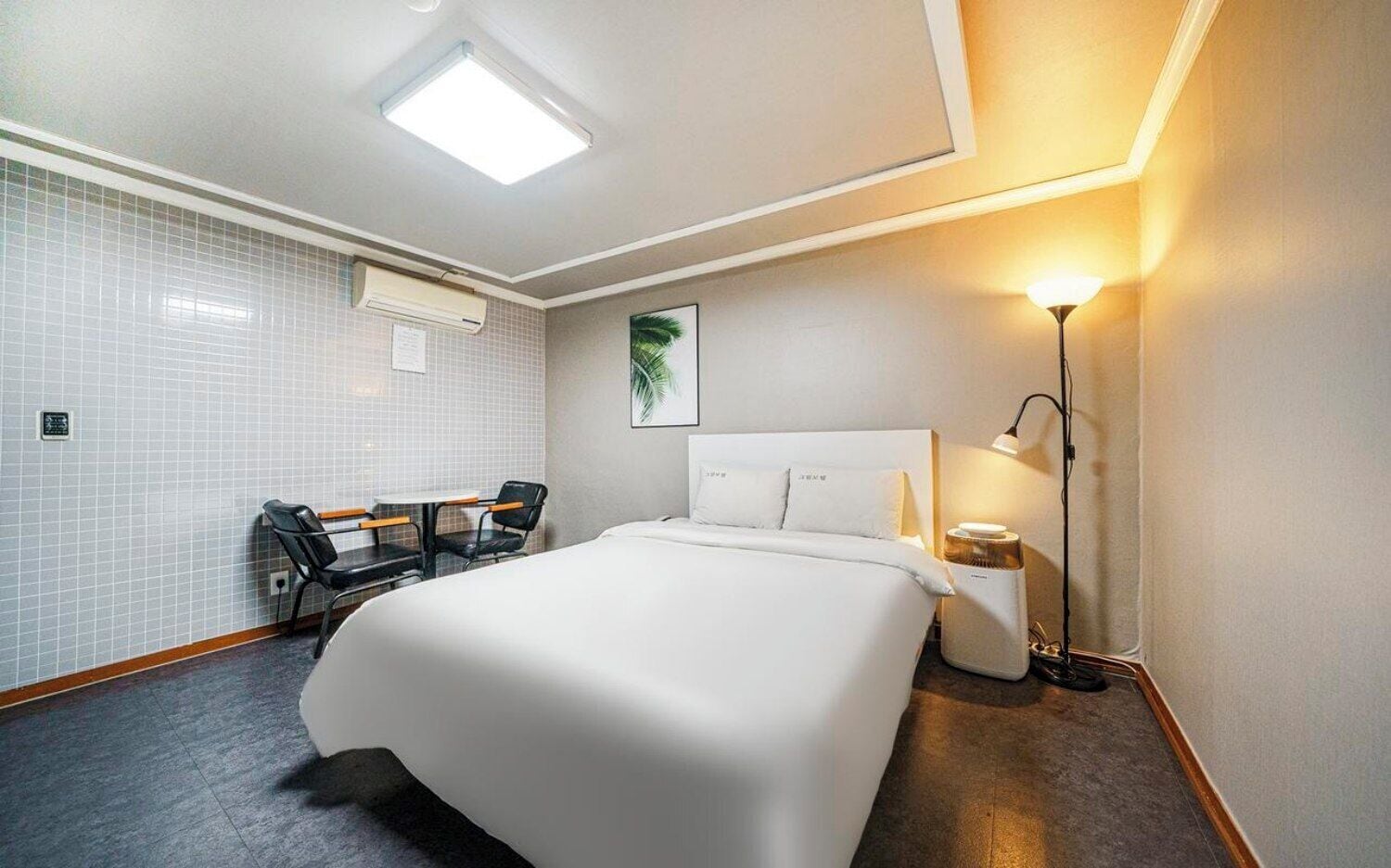 Habitación de hotel con una cama grande, un escritorio con dos sillas, aire acondicionado de pared, un cuadro enmarcado y una lámpara de noche.