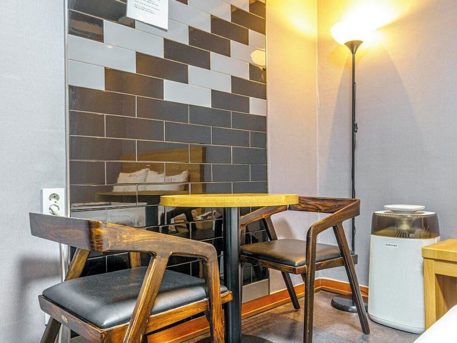 A small room with a wooden table and chairs, a lamp, a speaker, and a wall with a geometric tile pattern.