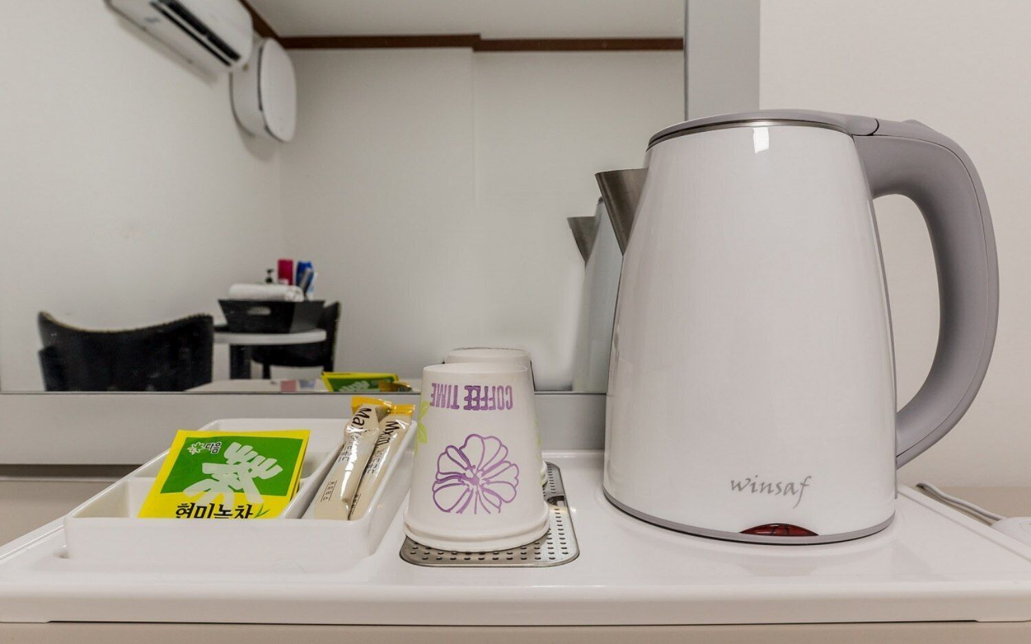 Una cafetera blanca con asa, una taza blanca con diseño de flor morada, y una bandeja con una caja verde y dos sobres de leche capuchina.