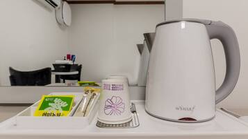 Una cafetera blanca con asa, una taza blanca con diseño de flor morada, y una bandeja con una caja verde y dos sobres de leche capuchina.