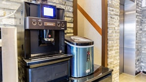 A coffee machine and a rice cooker on a countertop in a kitchen area with a brick wall and an elevator in the background.