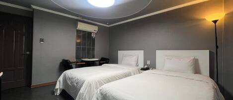 Une chambre d’hôtel avec deux lits, un bureau, une chaise, une table ronde, un climatiseur fixé au mur et un luminaire au plafond.