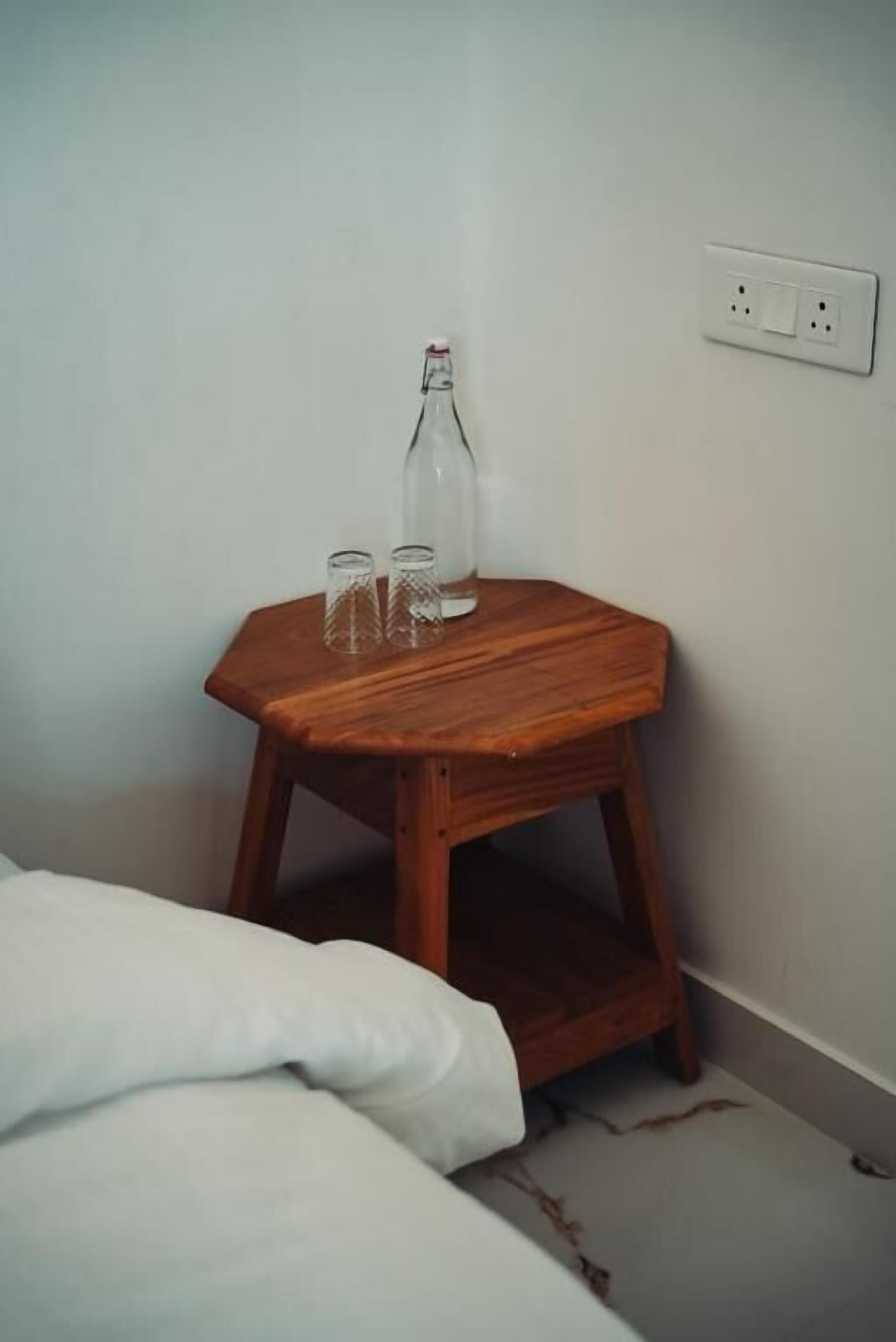 A small wooden bedside table with a bottle and two glasses against a plain wall.