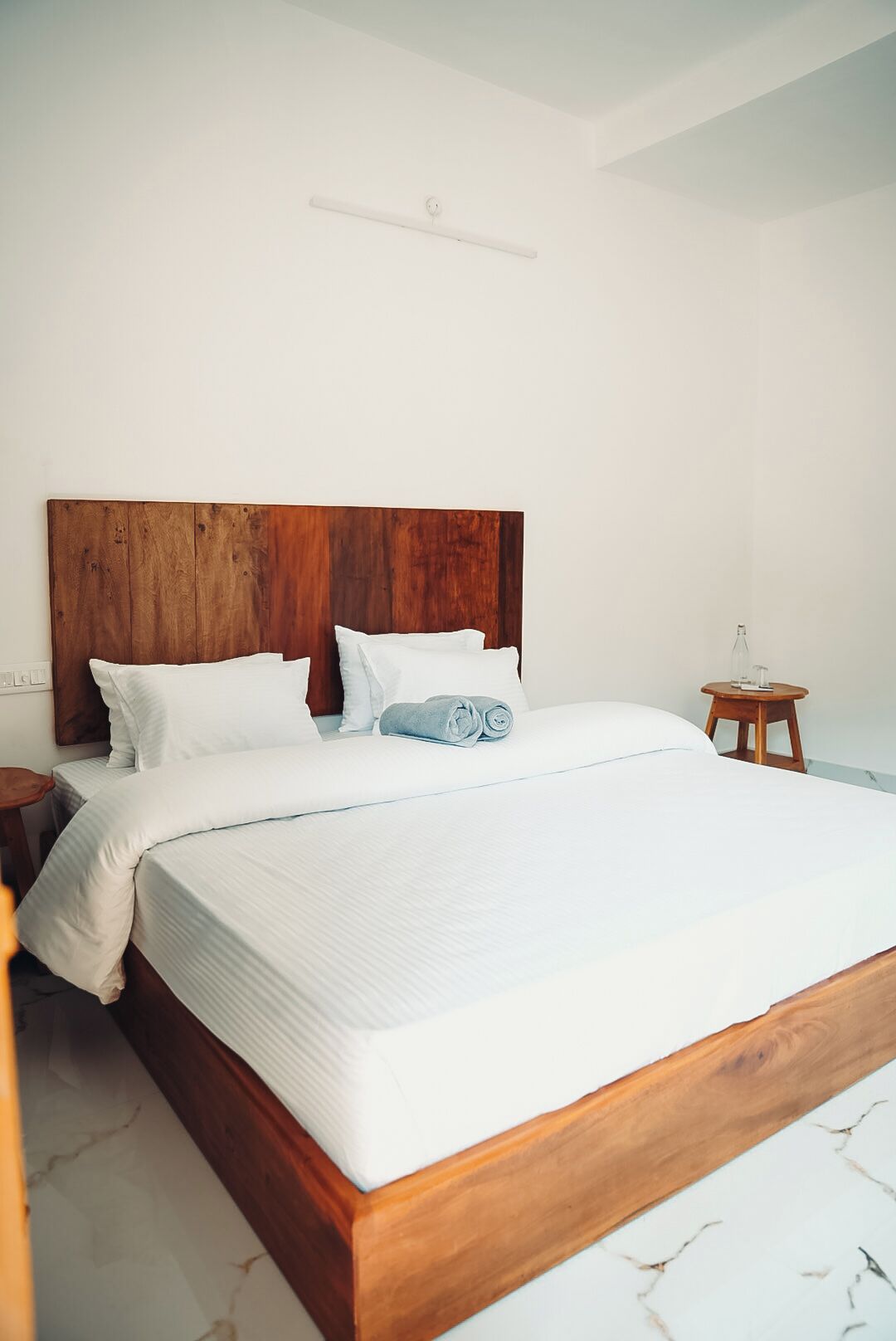 Quarto com cabeceira de madeira, cama grande com roupa de cama branca, mesa de cabeceira de madeira pequena e piso de mármore.