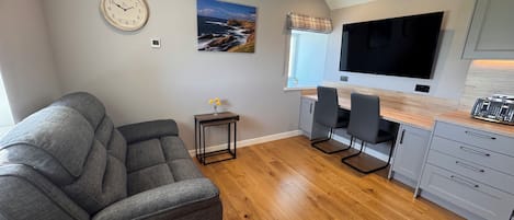 A modern kitchen with a dining area, a grey sofa, wooden flooring, and a large flat-screen TV.