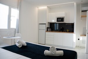 A modern hotel room with a bed, a small round table with two glasses, a dark blue bedspread, a neatly made white towel, a kitchenette with a microwave, a refrigerator, and a coffee maker. - AP Alsa 02 with Shared Terrace, WiFi, and Air Conditioning (Conil de la Frontera)