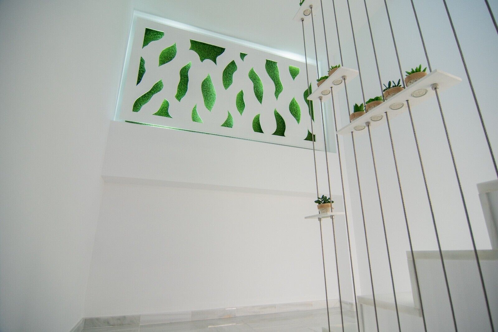A room with a white wall, a window with a green leafy pattern, and a white shelf with potted plants.