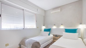 A hotel room with two beds, a window with blinds, a nightstand, and wall-mounted lights.